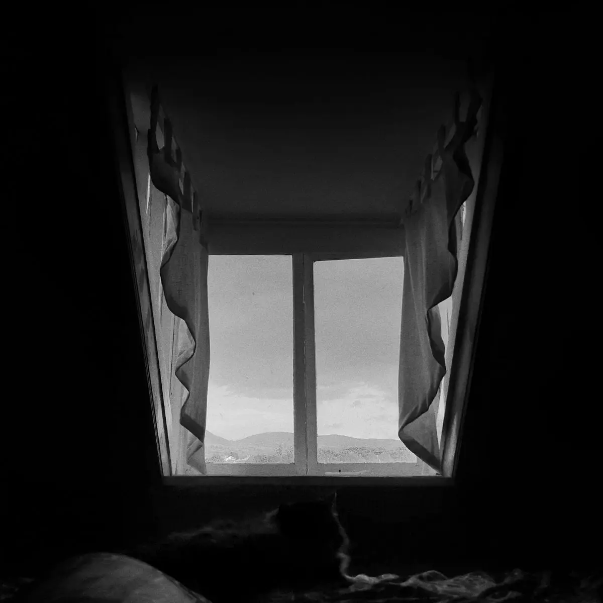 The Mourning After: a black and white attic room looking out through a window at distant hills.